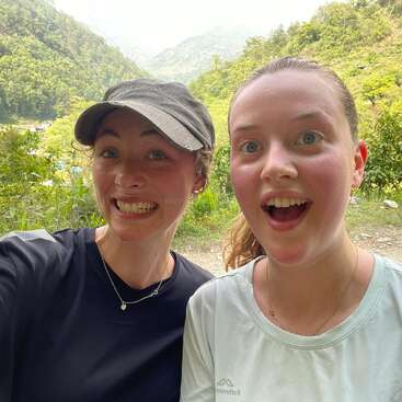 Deux jeunes femmes posent pour un selfie plein de joie en plein air, un large sourire aux lèvres. Derrière elles, des arbres verdoyants et des montagnes vallonnées forment un magnifique décor naturel.