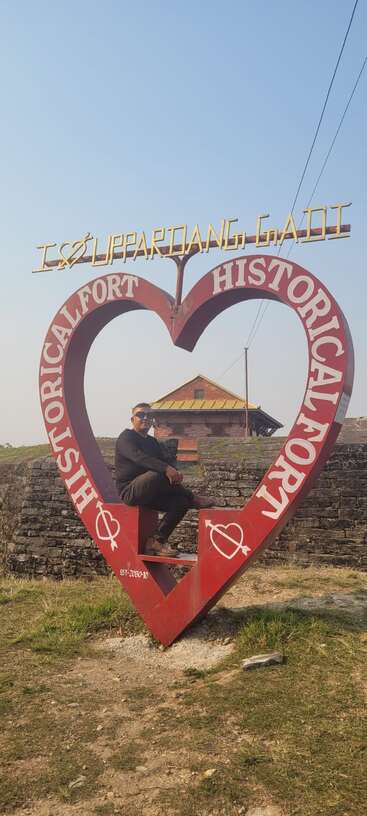 Un hombre está sentado dentro de un gran marco rojo con forma de corazón en el que se lee «Fuerte histórico». El cartel de arriba dice «I ♥ LIPPARDANG GADI». Escena al aire libre bajo un sol radiante.