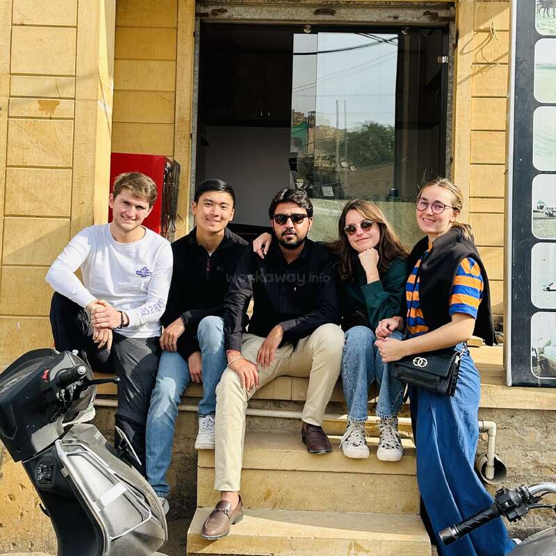 A imagem mostra quatro pessoas sentadas em degraus do lado de fora de um prédio, com uma motocicleta à esquerda e um pôster à direita, contra uma parede de tijolos amarelos.