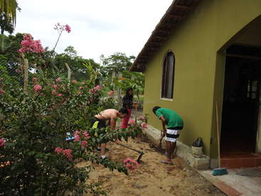 A imagem mostra três indivíduos trabalhando em um canteiro de jardim do lado de fora de uma casa, cercados por uma cerca e árvores, com um céu nublado ao fundo.