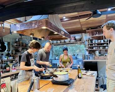Four people are cooking together in a cozy, rustic kitchen filled with shelves, dishes, and utensils. The atmosphere appears collaborative and creative, with natural light.