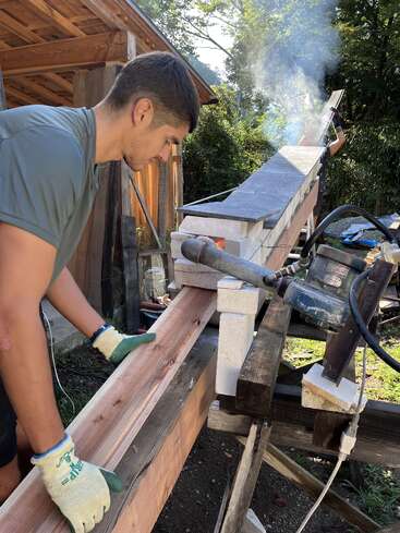 A man wearing gloves works with wooden beams near a homemade contraption emitting smoke, possibly a kiln or wood treatment device, outside a rustic wooden building.