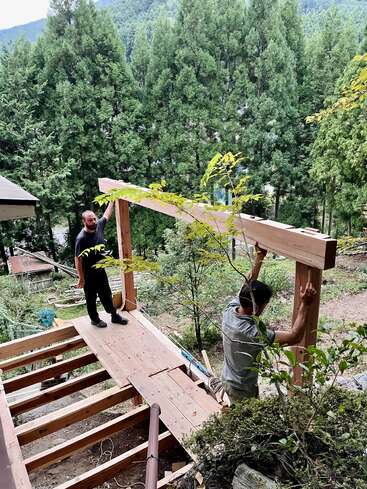 Two men are constructing a wooden structure outdoors, surrounded by dense, green forest. They are working on a platform together, with tools and boards nearby.