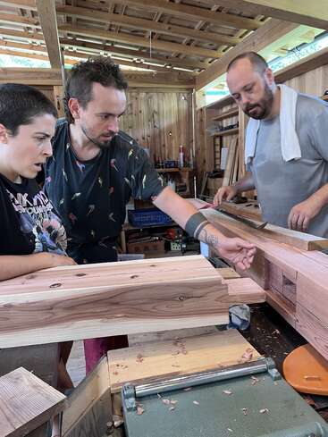 Three people are intently working together in a woodworking shop, examining a large wooden joint with concentration. Tools and wood shavings are visible on the workbench.