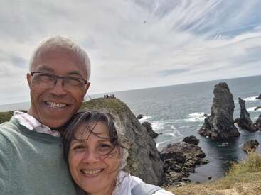 Una pareja sonriente se toma un selfie en un día ventoso cerca de espectaculares acantilados marinos y formaciones rocosas, con el océano y el cielo nublado de fondo.