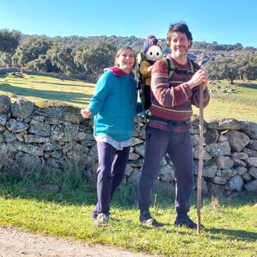 Ein lächelndes Paar posiert beim Wandern im Freien an einer Steinmauer. Der Mann trägt einen Rucksack mit einem süßen Stofftier. Grüne Felder und Bäume umgeben sie.