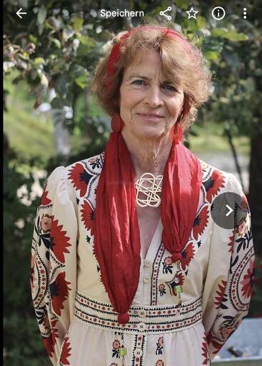 Eine Frau steht im Freien, trägt ein traditionelles besticktes Kleid mit roten und schwarzen Blumenmustern, einen roten Schal, passende Ohrringe und eine dekorative Halskette und lächelt freundlich.