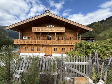 A beautiful wooden chalet with balconies and ornate trim sits amid greenery and hills, framed by a rustic wooden fence under a partly cloudy blue sky.