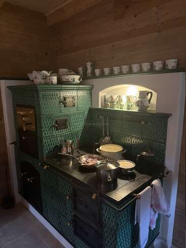 This cozy kitchen features a vintage, green-tiled wood stove, pots and pans cooking, wooden walls, and shelves displaying decorative porcelain mugs, jugs, and dishes above.