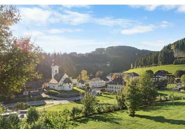 A picturesque village nestled in green hills, features charming white buildings, a central church with a steeple, surrounded by trees and bathed in sunlight.