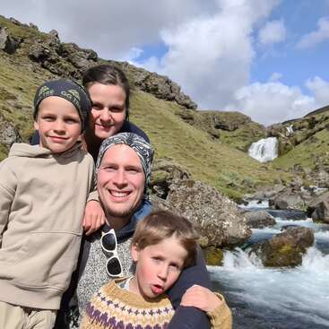 Une famille de quatre personnes souriante pose ensemble en plein air, avec en arrière-plan des collines verdoyantes, des rochers, un ruisseau et une cascade, sous un ciel partiellement nuageux.