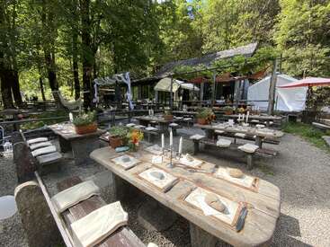 L'image représente une salle à manger extérieure sereine, avec des tables et des bancs en bois, entourée d'arbres luxuriants et d'un bâtiment rustique à l'arrière-plan.