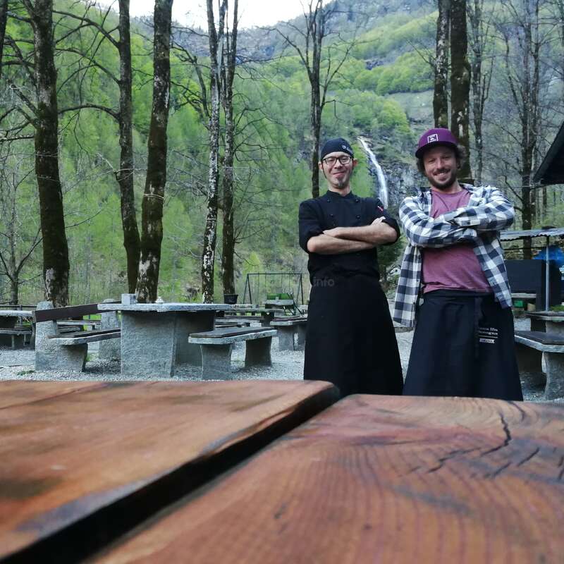 L'image représente deux hommes debout dans une zone boisée, avec une table en bois au premier plan et un bâtiment à droite. Les hommes portent des tabliers.