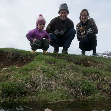 The image depicts a family of three crouching on a grassy hill beside a body of water, with a cloudy sky in the background.