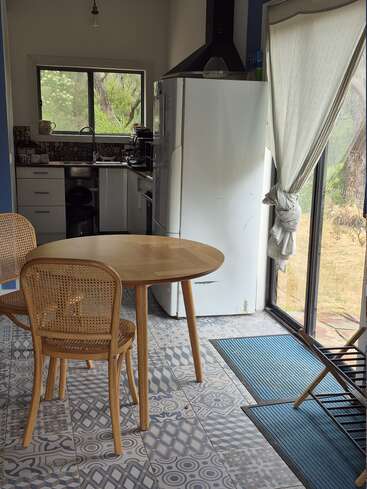 Eine gemütliche Küche mit gemustertem Fliesenboden, Holztisch und -stühlen, großem Kühlschrank, Fenster über der Spüle und Sonnenlicht, das durch Glastüren mit einem geknüpften Vorhang fällt.