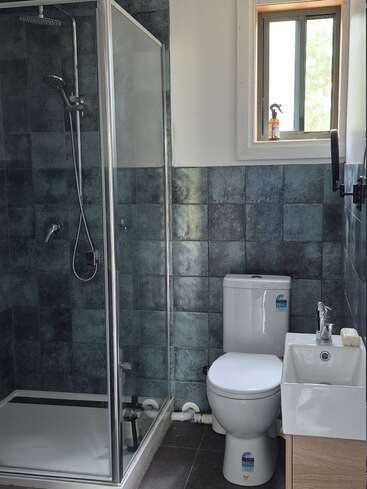 Dieses Badezimmer hat eine gläserne Duschkabine, geflieste Wände, eine weiße Toilette, ein kleines Waschbecken mit Seife, ein Fenster darüber und eine Flasche auf der Fensterbank.