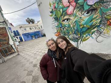 Duas pessoas tiram uma selfie numa praça charmosa e vazia, ao lado de um mural vibrante com um rosto colorido e folhoso com olhos hipnotizantes numa parede.