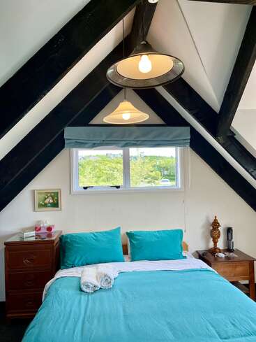 Um quarto aconchegante com roupa de cama turquesa, mesinhas de cabeceira de madeira, dois pendentes e uma janela com vista para a vegetação lá fora. O teto tem vigas de madeira pretas que chamam a atenção.
