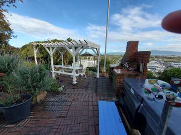 Eine Terrasse aus Ziegelsteinen mit einer weißen Pergola, einem Picknicktisch, Topfpflanzen, einer Feuerstelle und Blick auf die Stadt unter einem strahlend blauen Himmel mit vereinzelten Wolken.