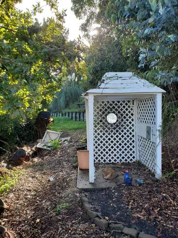 Ein kleiner weißer Gartenpavillon mit Gitterwänden steht inmitten von Bäumen und Grün. Die Sonne scheint hindurch und lässt einen Spiegel, verstreute Gartensachen und heruntergefallene Äste in der Nähe im Licht erstrahlen.