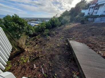 Ein abfallender, freigeräumter Hof mit spärlicher Vegetation, gesäumt von Bäumen und Sträuchern, mit Blick auf eine weit entfernte Stadt und Felder. Ein Holzsteg führt an einem Haus entlang.