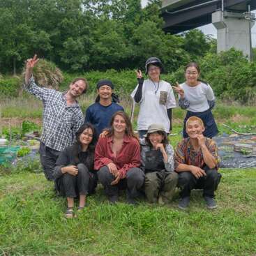 Eight people pose happily in a green outdoor setting, smiling and making peace signs. They appear to be friends or coworkers, possibly enjoying a day of gardening.
