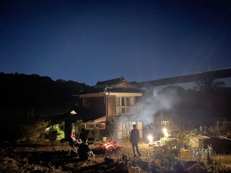 La imagen representa una serena escena nocturna de gente reunida en torno a una hoguera frente a una casa, rodeada de árboles y un cielo despejado.