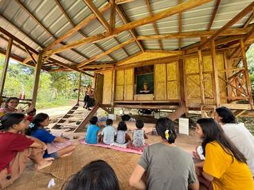 A group of children sit attentively on mats under a wooden shelter, listening to a teacher in front of a traditional bamboo house in a lush setting.