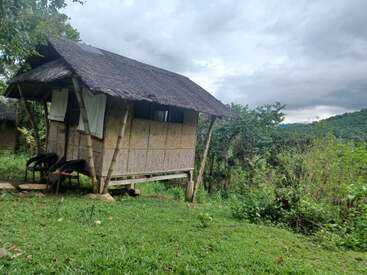 A small bamboo hut with a thatched roof stands on stilts, surrounded by lush greenery, overlooking a scenic valley and distant forested hills under cloudy skies.