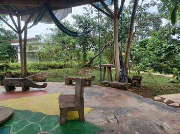 This image shows an outdoor seating area with animal-shaped benches, a hammock, a swing, trees, greenery, and a colorful, painted concrete floor beneath a wooden roof.