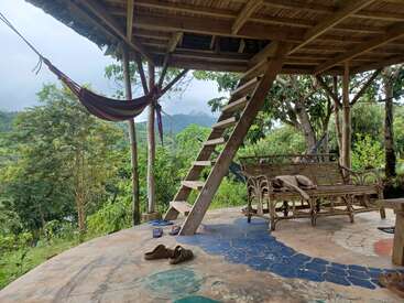 A rustic outdoor patio features a wooden bench, resting dog, slippers, hammocks, wooden stairs, and lush green mountain scenery, creating a peaceful, relaxing, natural atmosphere.