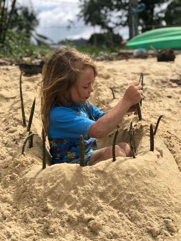 A young child sits in the sand at the beach, building and decorating a sand structure with sticks, fully immersed in creative and imaginative outdoor play.