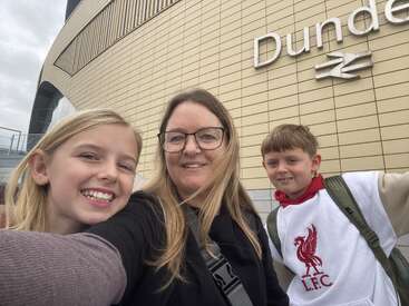 Eine Frau und zwei Kinder lächeln für ein Selfie vor dem Bahnhof von Dundee. Das Gebäude hat eine moderne Architektur und ein Schild mit großen Buchstaben, auf dem "Dundee" steht.