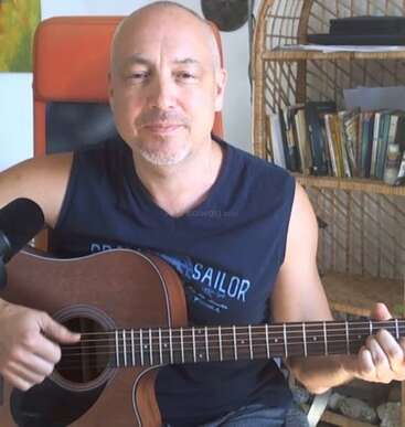 Un homme est assis à l'intérieur et joue de la guitare acoustique. Il est souriant et porte une chemise sans manches. Derrière lui se trouve une étagère avec des livres et des papiers.