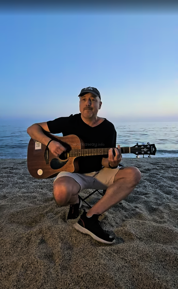 Un homme est assis sur une plage de sable au crépuscule, coiffé d'une casquette et vêtu de vêtements décontractés ; il joue sereinement de la guitare acoustique, avec l'océan paisible en toile de fond.