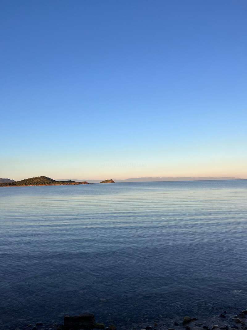 Un mar en calma refleja el cielo azul y despejado, con colinas lejanas y pequeñas islas en el horizonte. Las suaves olas rompen contra la costa rocosa al atardecer.
