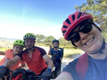 La imagen muestra a una familia de cuatro miembros con casco y montando en bicicleta en un día soleado, con árboles y un cielo azul de fondo.