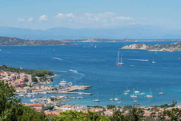 This image showcases a beautiful coastal town with a marina, several boats and yachts on deep blue water, surrounded by hills and distant mountains under a clear sky.