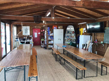 Cette image montre une pièce rustique en bois avec de longues tables et des bancs, une porte rouge, des étagères, un réfrigérateur, une télévision et des fournitures, suggérant un espace de rassemblement communautaire.