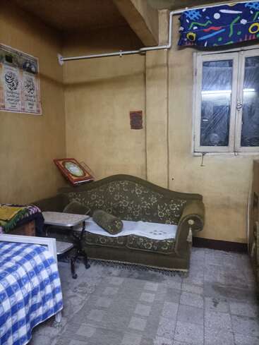 Ein bescheidener Raum mit vergilbten Wänden, einem olivgrünen, geblümten Sofa, einem kleinen Bett mit blau kariertem Bezug, zwei Tischen, einem Fenster und religiöser Dekoration.