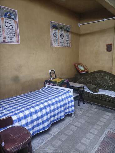 Ein einfaches Zimmer mit einem Einzelbett, das mit einem blau-karierten Laken bezogen ist, einem grünen Sofa, Wandpostern mit arabischen Texten, einem Holzstuhl und einem Gebetsteppich auf dem Boden.