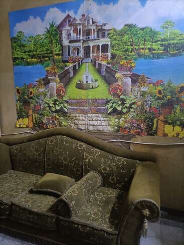 Eine olivgrüne Couch mit Blumenmuster steht an einer Wand. Darüber hängt ein farbenfrohes Gemälde mit einem Herrenhaus, einem Brunnen, einem Garten und einer Seelandschaft.