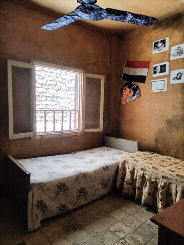 Une petite chambre rustique avec deux lits simples, une fenêtre ouverte, un ventilateur de plafond, un drapeau égyptien, des dessins d'enfants et des affiches accrochés au mur.