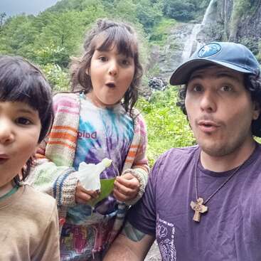 A man and two children pose with surprised expressions in front of a lush, green forest and a waterfall, enjoying an outdoor adventure together.