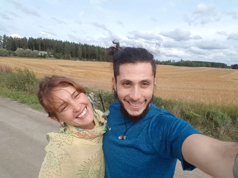 The image depicts a young couple taking a selfie on a dirt road, with a field of wheat and trees in the background under a cloudy sky.