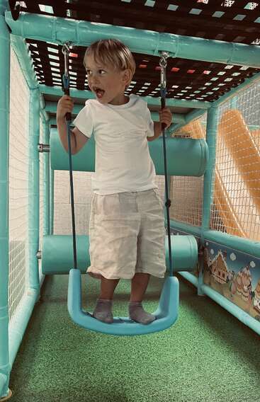 Ein kleiner Junge in einem weißen T-Shirt und Shorts steht aufgeregt auf einer Schaukel in einem Indoor-Spielbereich mit bunten Geräten und Netzwänden.