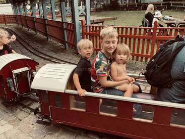 Drei Kinder fahren mit einem kleinen Zug in einem Vergnügungspark. Sie wirken glücklich und entspannt, umgeben von roten Zäunen und Schienen, und genießen einen lustigen Familienausflug.