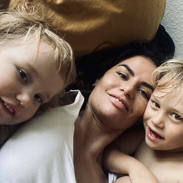 Una mujer se tumba sonriente con dos niños pequeños, muy juntos. El grupo parece feliz y relajado, compartiendo un momento cariñoso, franco y acogedor.