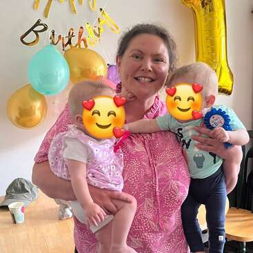 A joyful woman in a pink shirt holds two small children with emoji faces at a birthday party, surrounded by colorful balloons and a golden number one balloon.