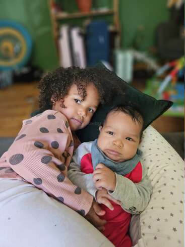 Uma menina abraça carinhosamente seu irmãozinho em uma almofada aconchegante, ambos com cabelos cacheados, expressões relaxadas e calor em um quarto colorido e acolhedor.